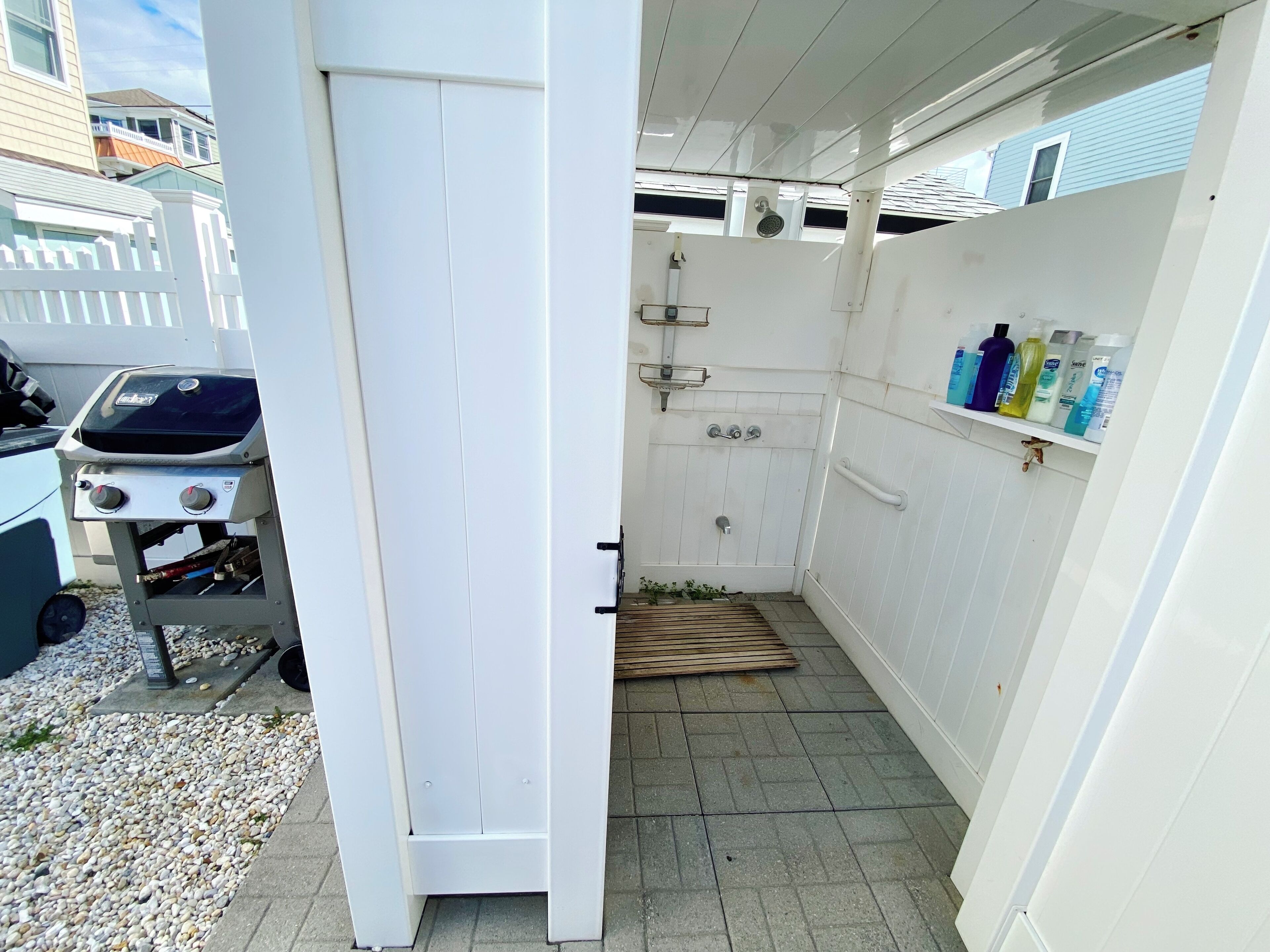A narrow outdoor space with a white door, a small shower, and a shelf with cleaning supplies.