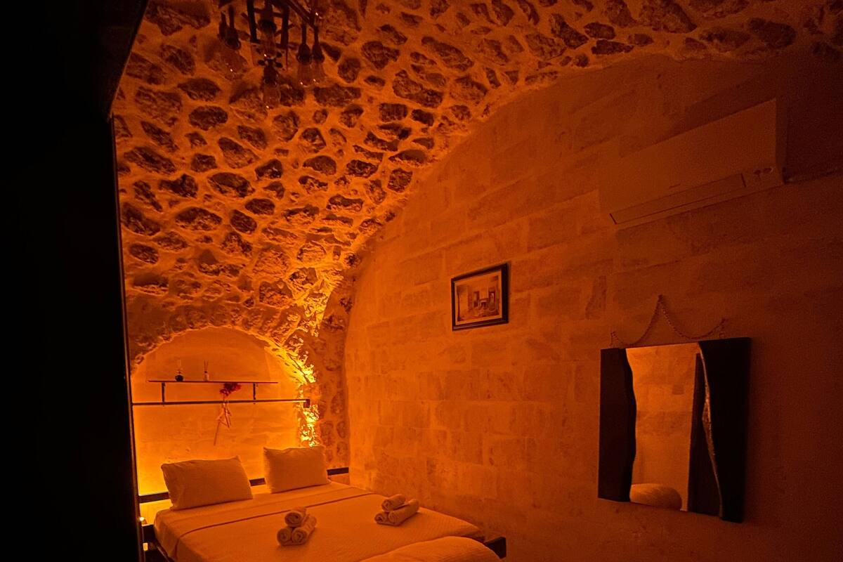 A dimly lit room with a stone ceiling, a bed with pillows, a bench, and a framed picture on the wall.