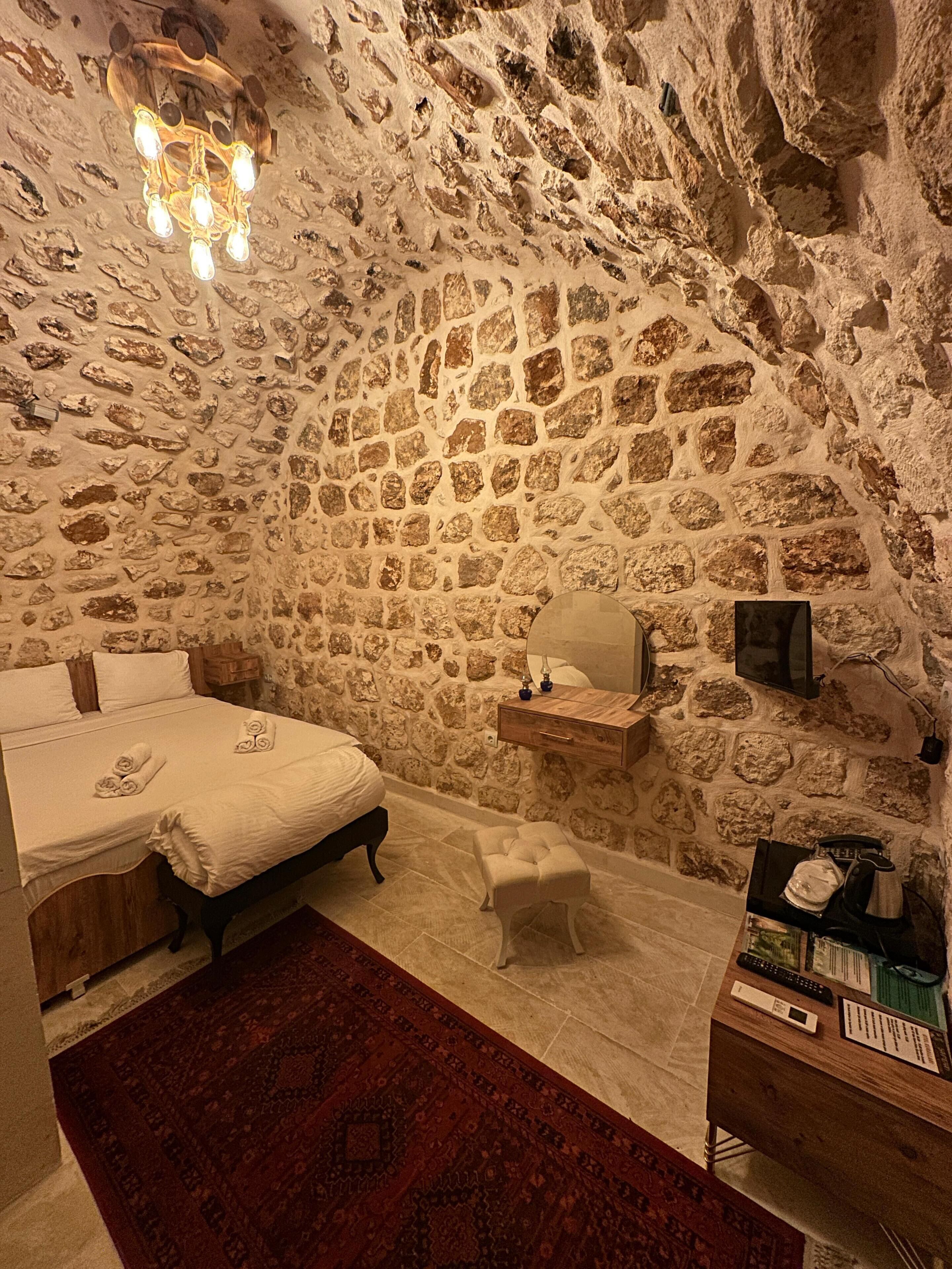 Un dormitorio con pared de piedra, una cama, una mesita de noche, una lámpara, una pequeña mesa y una alfombra.