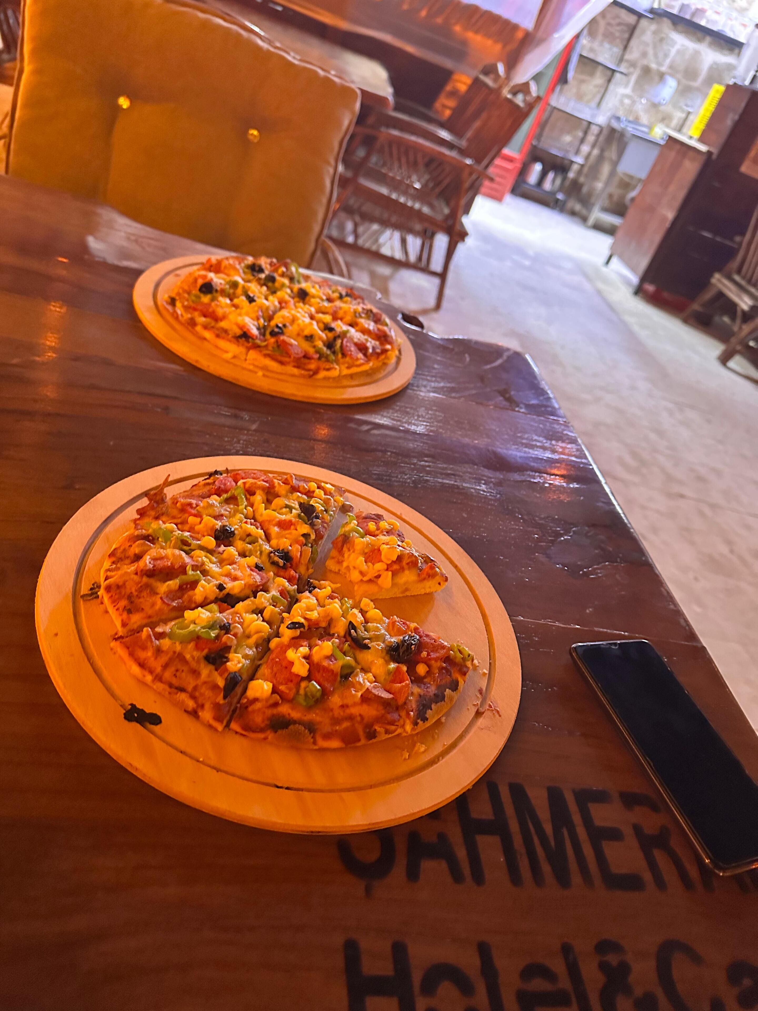 A wooden table with two pizza slices on wooden plates, a smartphone on the table, and a bar area in the background.