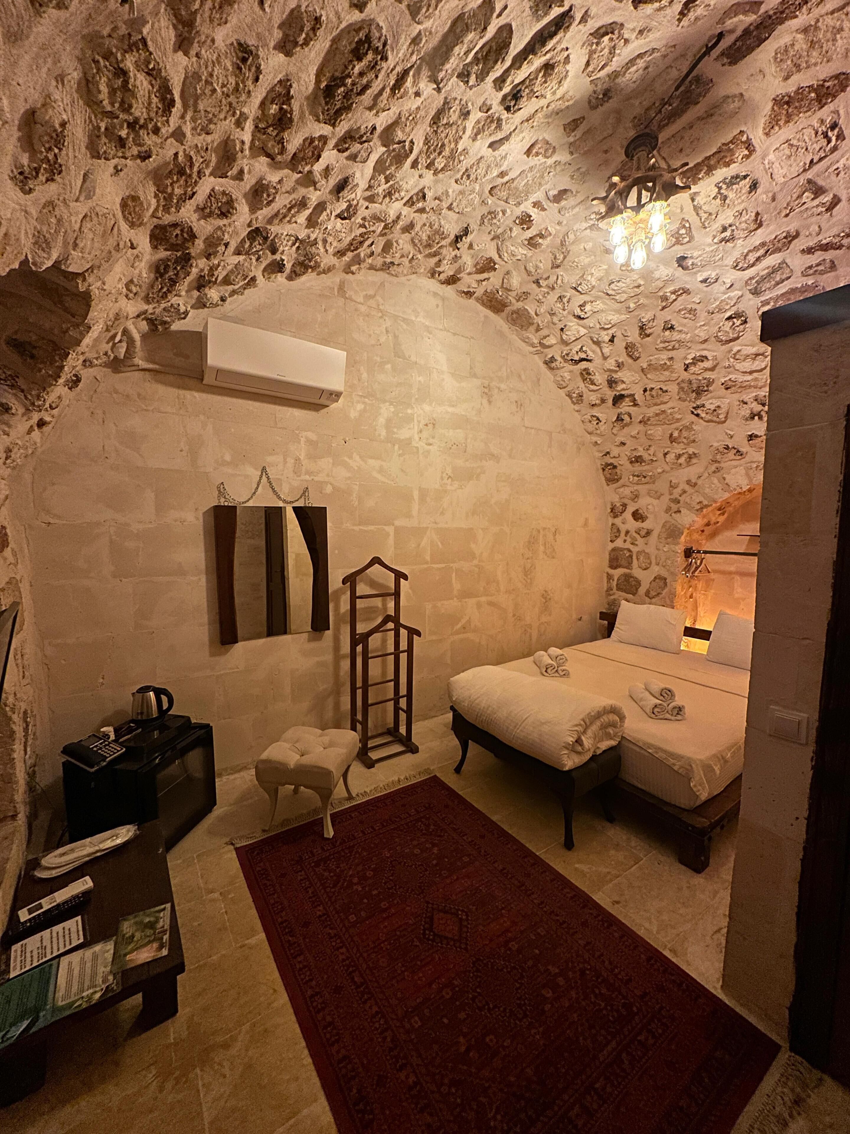 A cozy room with a stone wall, a bed, a small table with a kettle, and a chair.