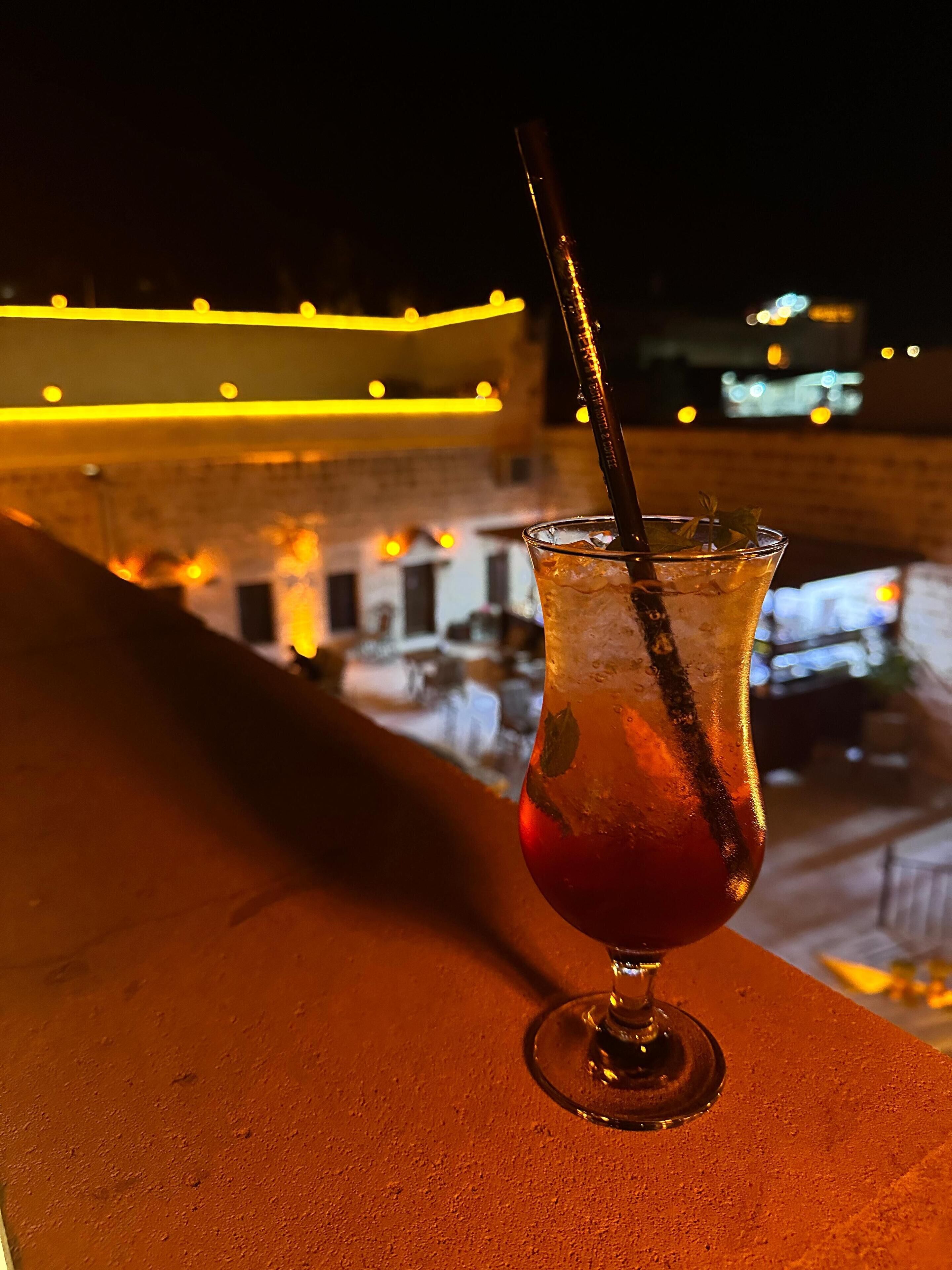 Um copo de cocktail com canudo numa varanda com uma vista urbana ao fundo.