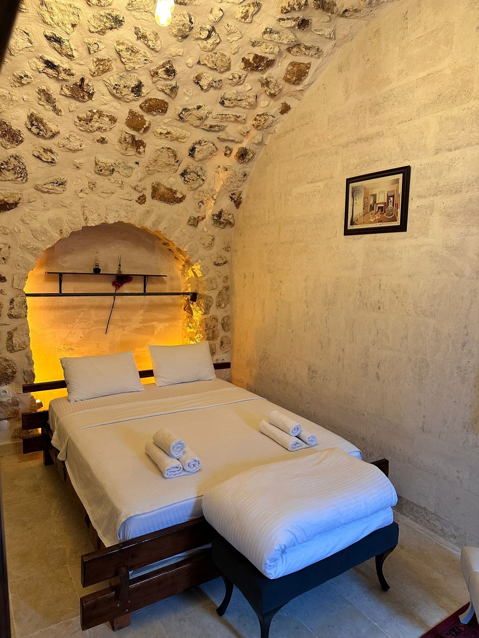 Un dormitorio con pared de piedra, una cama con ropa blanca, una mesita de noche y un cuadro enmarcado en la pared.