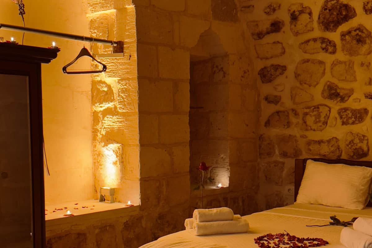 A cozy room with a stone wall, a bed with white linens, a wooden bench, and a table with candles and rose petals.