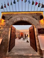 A stone archway with two wooden doors, leading to a courtyard with seating and a signboard listing amenities.