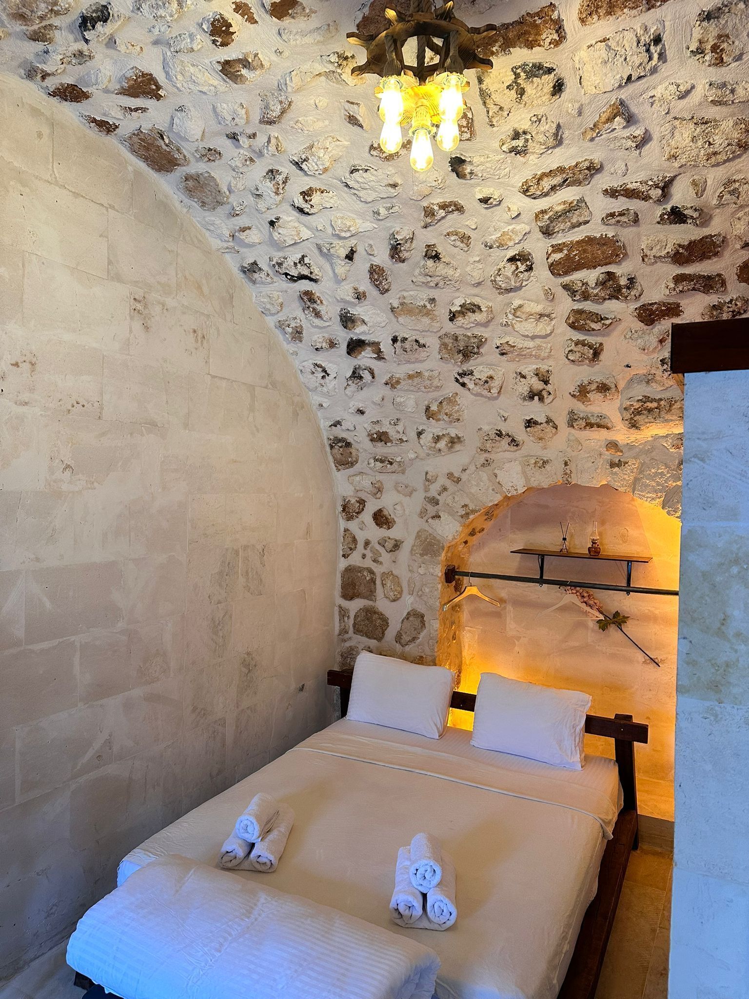 Un dormitorio con pared de piedra, cama con sábanas blancas y una lámpara colgante.