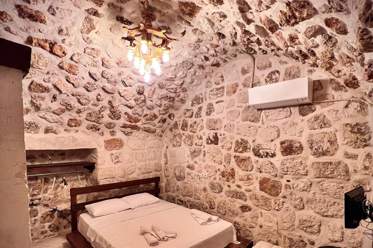 A stone-walled bedroom with a wooden bed, a red rug, and a small white stool.