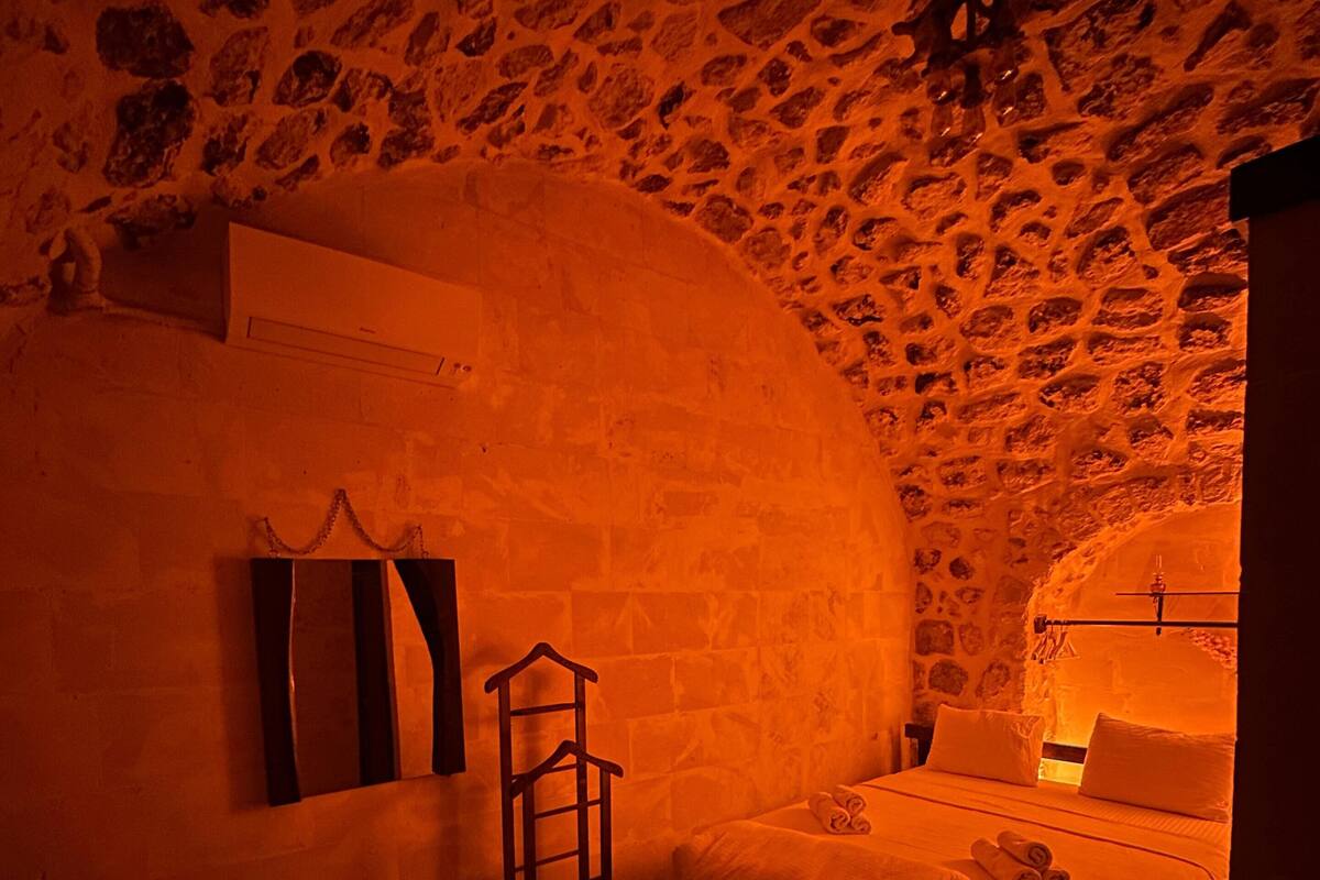 A cozy bedroom with a stone wall, a bed with pillows, a nightstand, and a small table with a lamp.