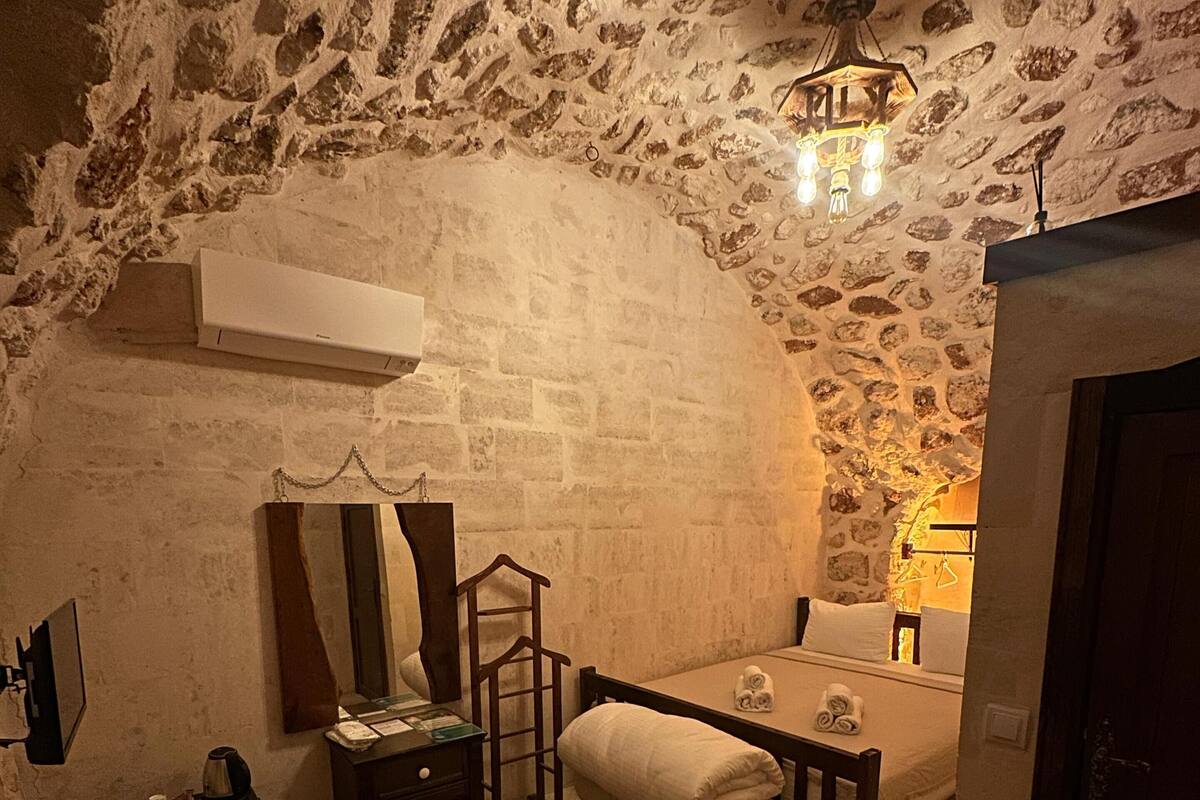 A room with a stone wall, a bed, a bedside table, a chair, a small table, a mirror, a heater, a telephone, and a door.