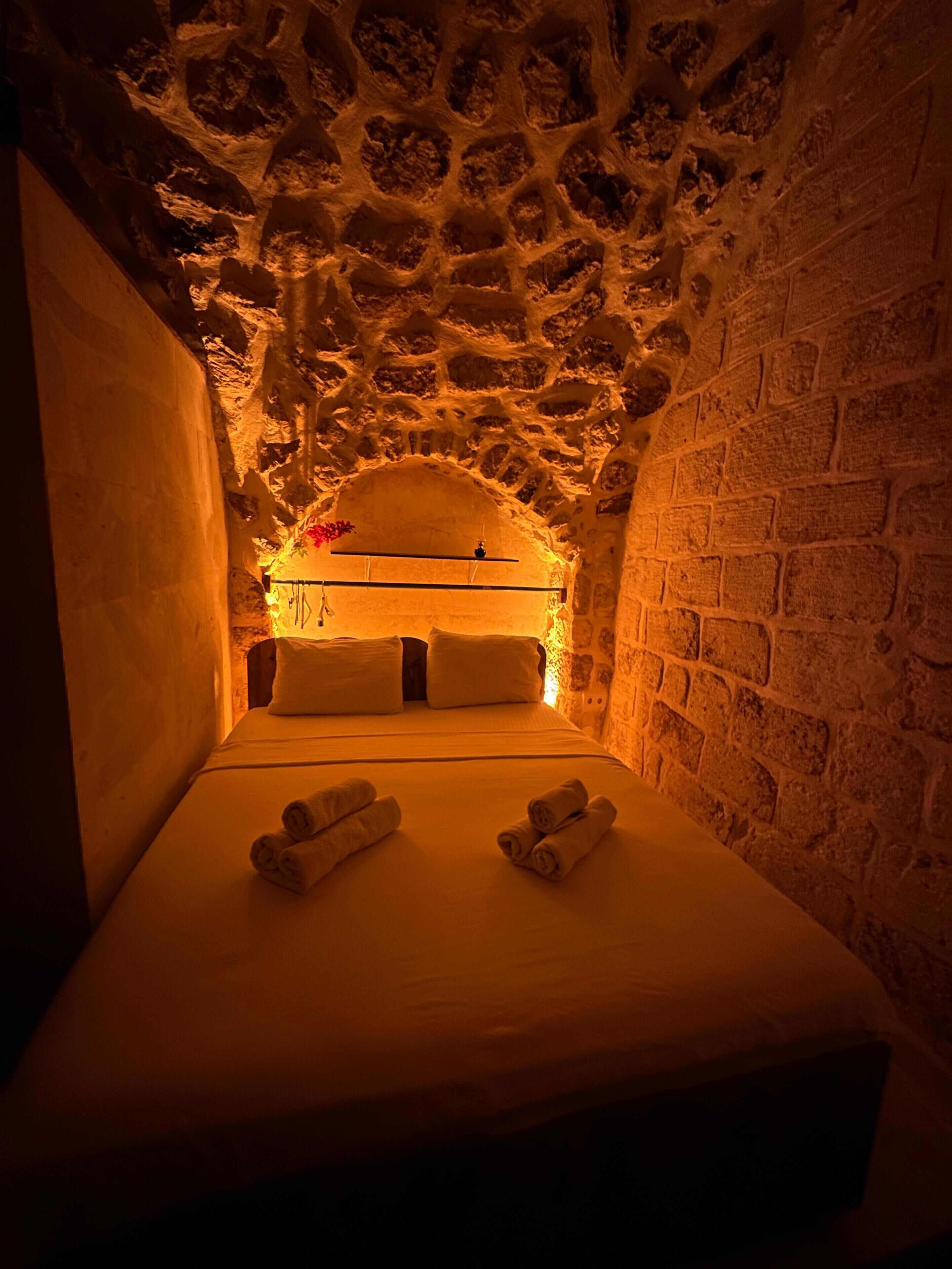 A narrow, stone-walled room with a bed, towels, and a small table with a lamp.