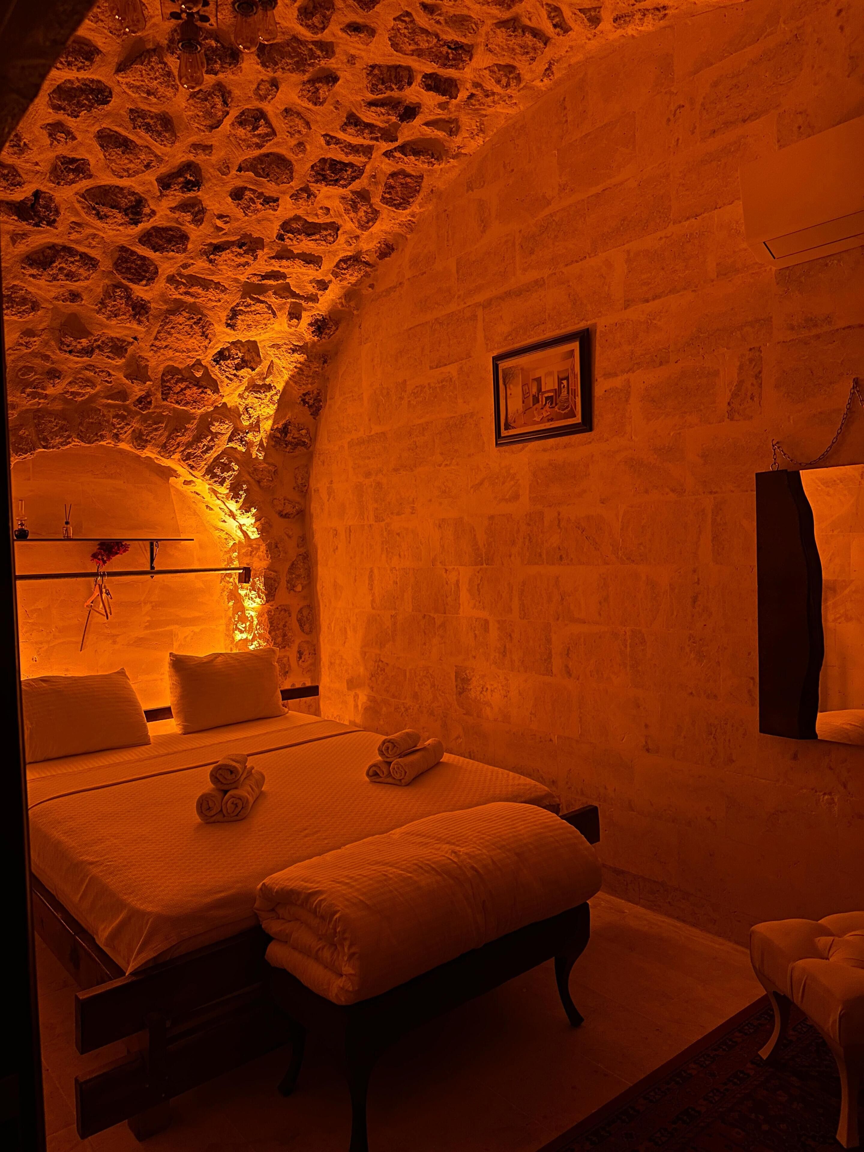 A room with a stone ceiling and walls, a bed with white linens, a bedside table, and a chair.