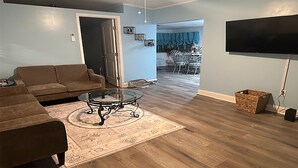 A living room with a brown sofa, a glass coffee table, and a patterned rug. There is a TV mounted on the wall and a wicker basket on the floor.