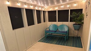 A small, enclosed balcony with a bench, string lights, and a potted plant.