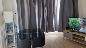 A room with a large window, grey curtains, a black chair, a TV on a wooden stand, and a blue patterned shelf. - A lovel one bedroom.  (London)