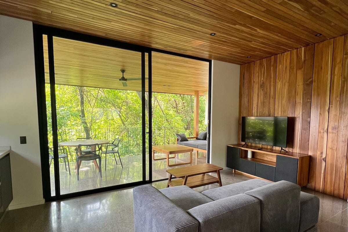 A modern living room with a large sectional sofa, a wooden ceiling, and a large sliding glass door leading to an outdoor patio.