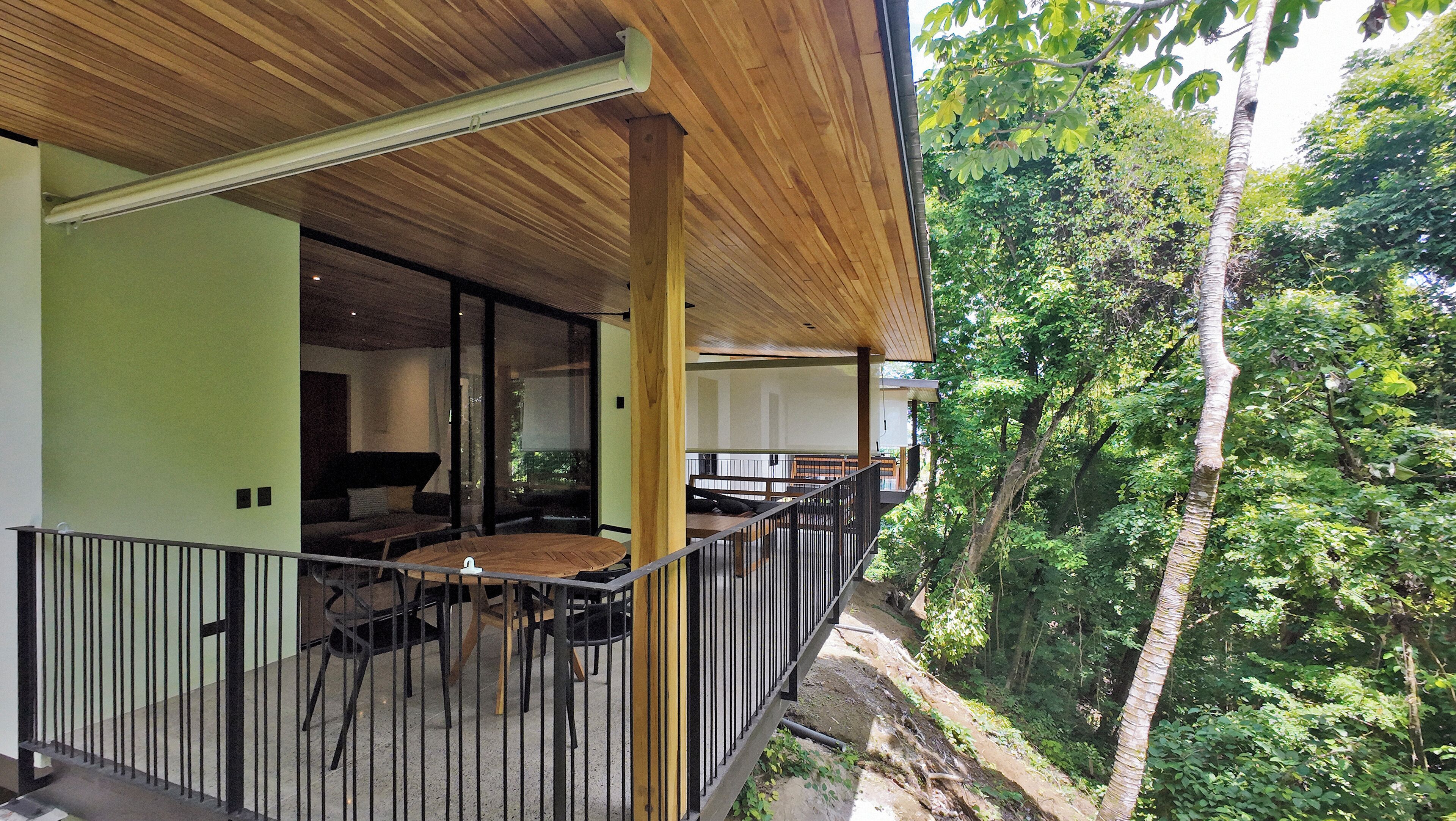 A wooden balcony with a dining area, a view of lush greenery, and a modern building structure.