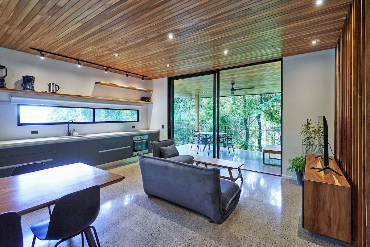 A modern kitchen with a wooden ceiling, a dining area with a table and chairs, and a large window offering a view of greenery.