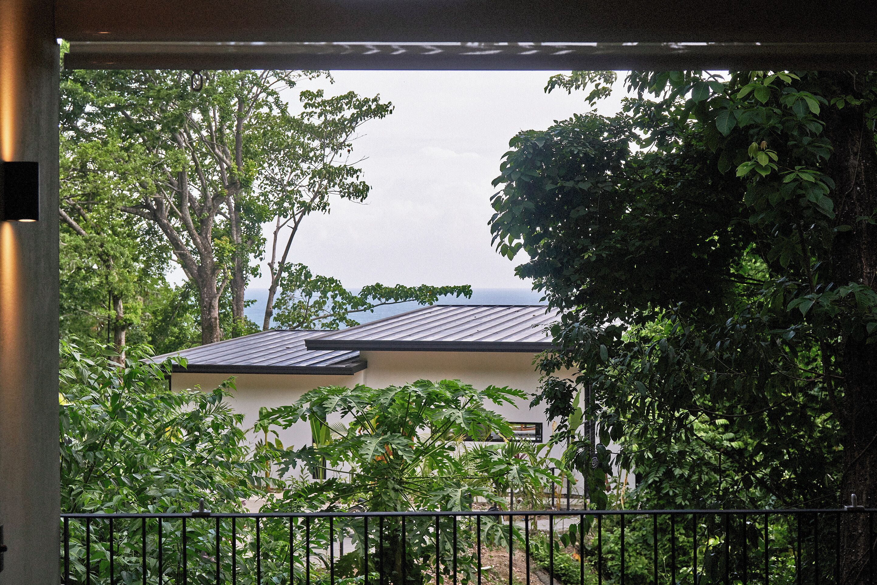 A view through a window with a metal railing, overlooking a building with a sloped roof and surrounded by lush greenery.
