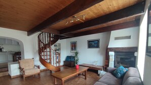 A spacious living room with a wooden staircase, a fireplace, and a kitchen in the background. - Maison Bretonne à la Pointe du Raz (Plogoff)