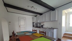 A dining area with a wooden table and chairs, a kitchen with white cabinets, and a tiled floor. - Maison Bretonne à la Pointe du Raz (Plogoff)
