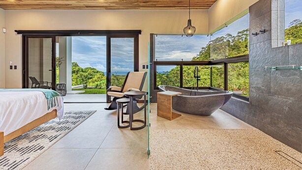 A modern bedroom with a large bed, a sitting area with a chair and table, a bathtub, and a view of greenery through large windows.