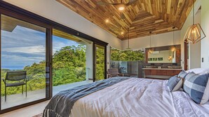 A bedroom with a large bed, a view of lush greenery, and a modern kitchen area.