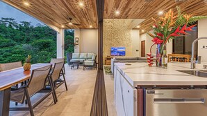 A modern kitchen with a large island, a dining area with a wooden table and chairs, and a view of lush greenery outside.