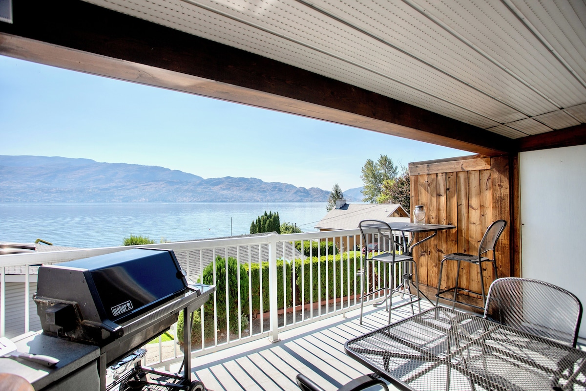 A balcony with a grill, chairs, and a view of a lake and mountains.