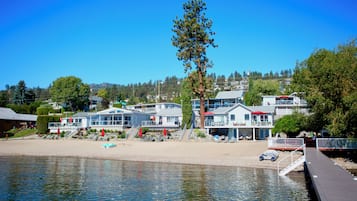 Waterfront property with modern buildings, a tree, and a wooden dock along a sandy beach.