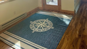 A room with a wooden floor, a large window with a view of a deck, a door with a sign above it, and a decorative wall piece.