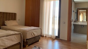 A bedroom with two beds, a wooden wardrobe, a window with sheer curtains, and a bathroom visible through an open door.
