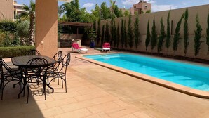 A swimming pool with a tiled deck, surrounded by a low wall and tall, narrow trees.
