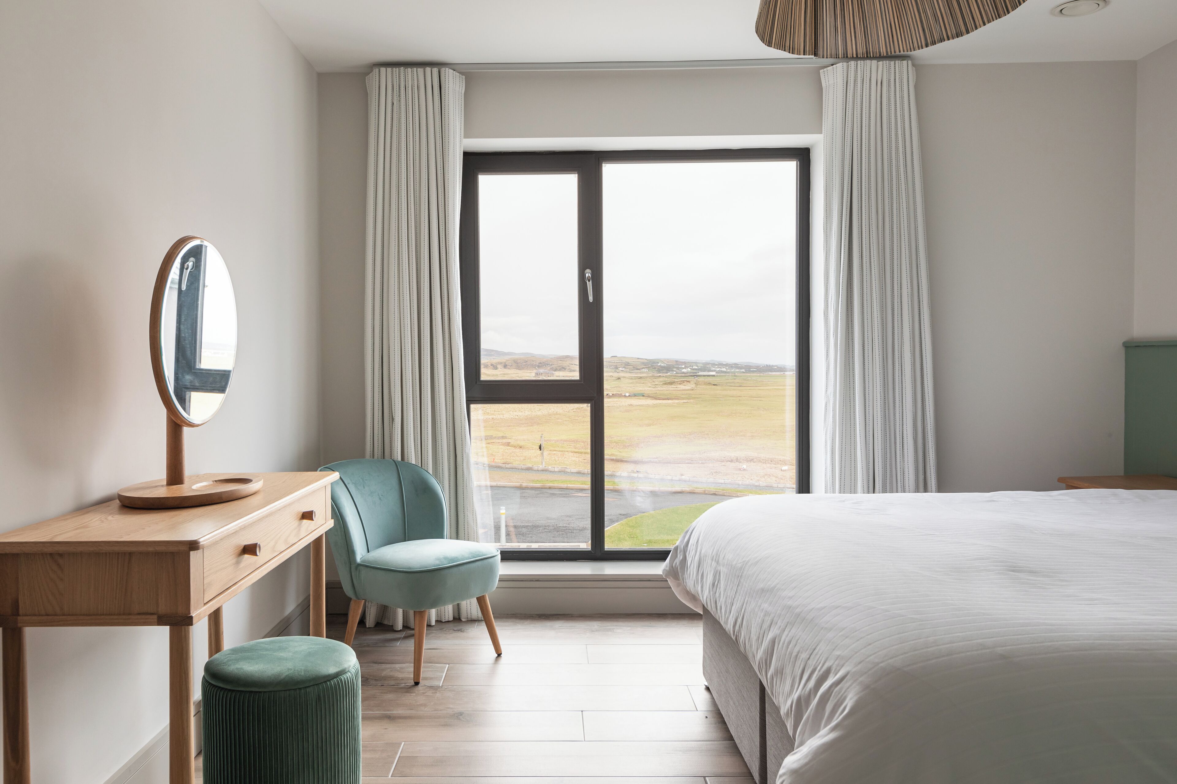 A bedroom with a large bed, a wooden desk with a mirror, a chair, and a view of a landscape through a sliding door.