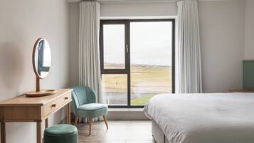 A bedroom with a large bed, a wooden desk with a mirror, a chair, and a view of a landscape through a sliding door.