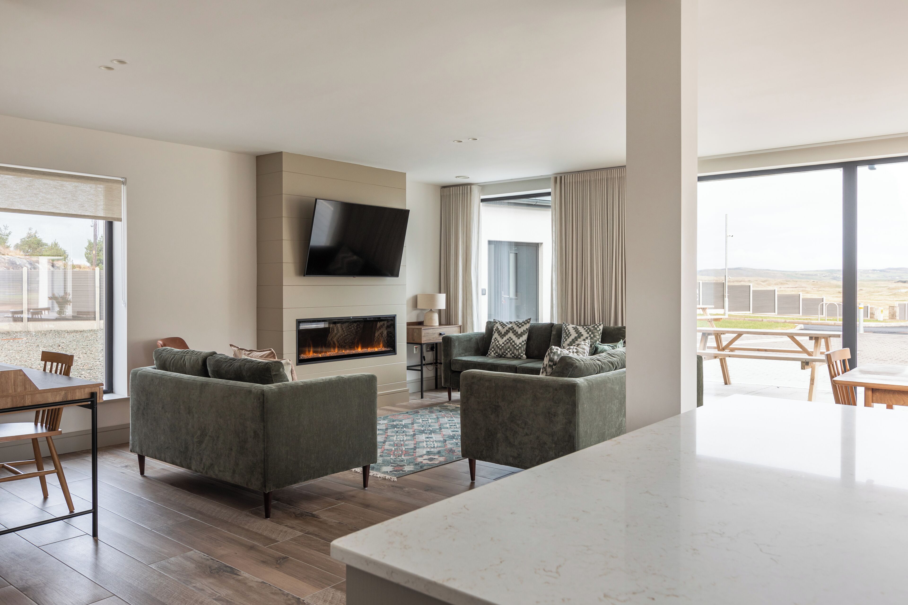 A modern living room with a fireplace, a flat-screen TV mounted on the wall, and a large window offering a view of the outdoors.