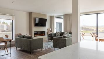 A modern living room with a fireplace, a flat-screen TV mounted on the wall, and a large window offering a view of the outdoors.