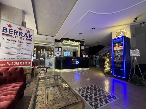 A hotel lobby with a reception desk, a red sofa, a coffee table with a decorative glass top, and a blue neon sign. - Berrak Otel (Salihli)