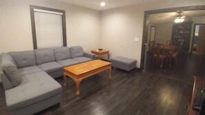 Una sala de estar con un sofá seccional gris, una mesa de centro de madera y un pequeño otoman. Se ve un área de comedor con una mesa y sillas a través de un umbral abierto.
