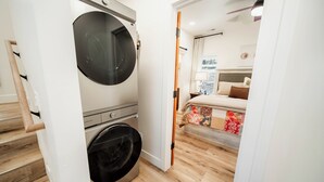 Habitación de hotel compacta con lavadora, una cama con colcha colorida y suelo de madera.