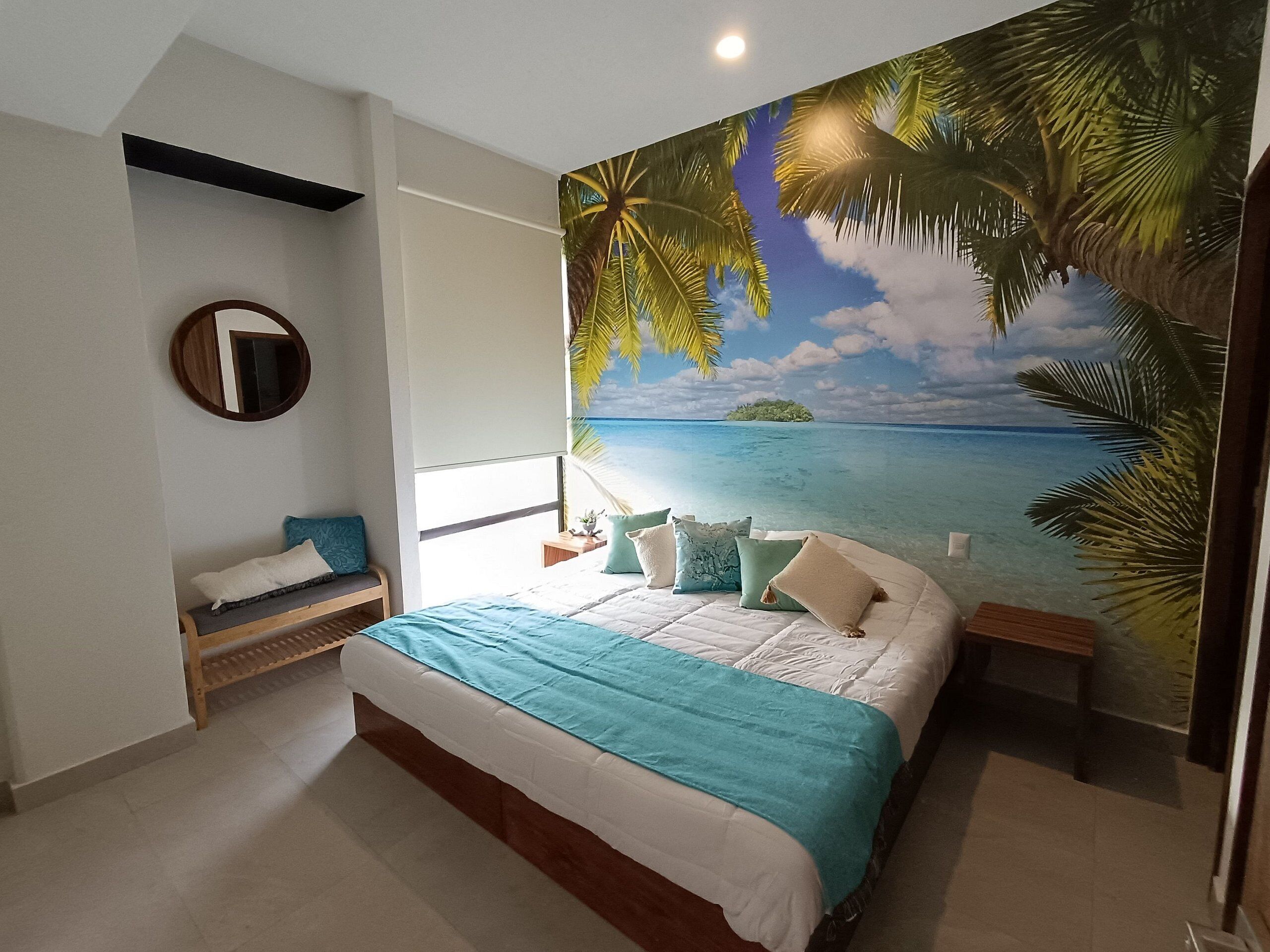 Une chambre avec un lit, un banc, un miroir rond et une fresque murale aux motifs tropicaux.