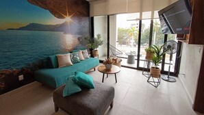 A modern living room with a large mural of a coastal scene, a teal sofa, a grey ottoman, and a flat-screen TV mounted on the wall.