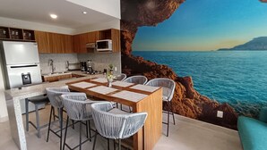 A modern kitchen with a central island, bar stools, and a view of the sea through a rock formation.