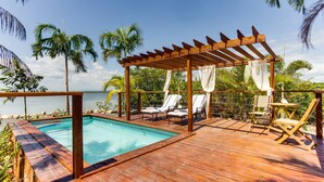 A wooden deck with a pool, lounge chairs, and a pergola with white curtains. - Luxurious Lagoon Home w/ Elegant Decor & Pool (Placencia)