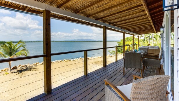 A wooden deck with a view of the beach and water, featuring wicker furniture and a railing. - Luxurious Lagoon Home w/ Elegant Decor & Pool (Placencia)