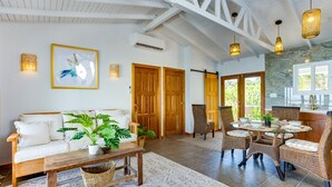 A spacious living area with a white sofa, wicker chairs, a wooden coffee table, and a dining table with chairs. There are wooden doors, a framed picture on the wall, and a stone wall in the background. - Luxurious Lagoon Home w/ Elegant Decor & Pool (Placencia)