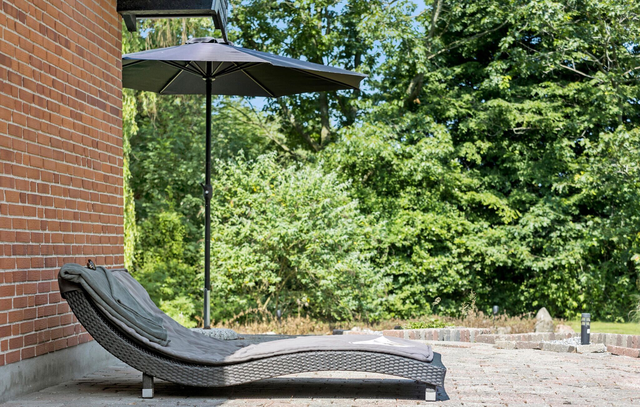 Eine Liege mit Metallgestell und Gewebesitz, auf einer gepflasterten Terrasse mit Backsteinmauer und einem großen Sonnenschirm, der Schatten spendet.