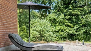 A sun lounger with a metal frame and a woven seat, positioned on a paved patio area with a brick wall and a large umbrella providing shade.