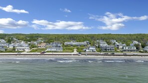 A coastal residential area with houses along the beachfront, a road with vehicles, and a forested hill in the background.