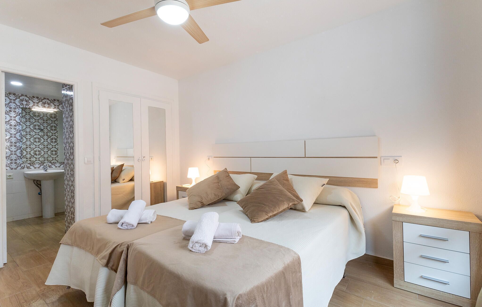 A hotel room with a bed, bedside tables, a ceiling fan, and a bathroom with a sink and mirror.