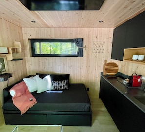 A cozy room with a bed, a window, a small kitchen area, and a shelf with mugs.